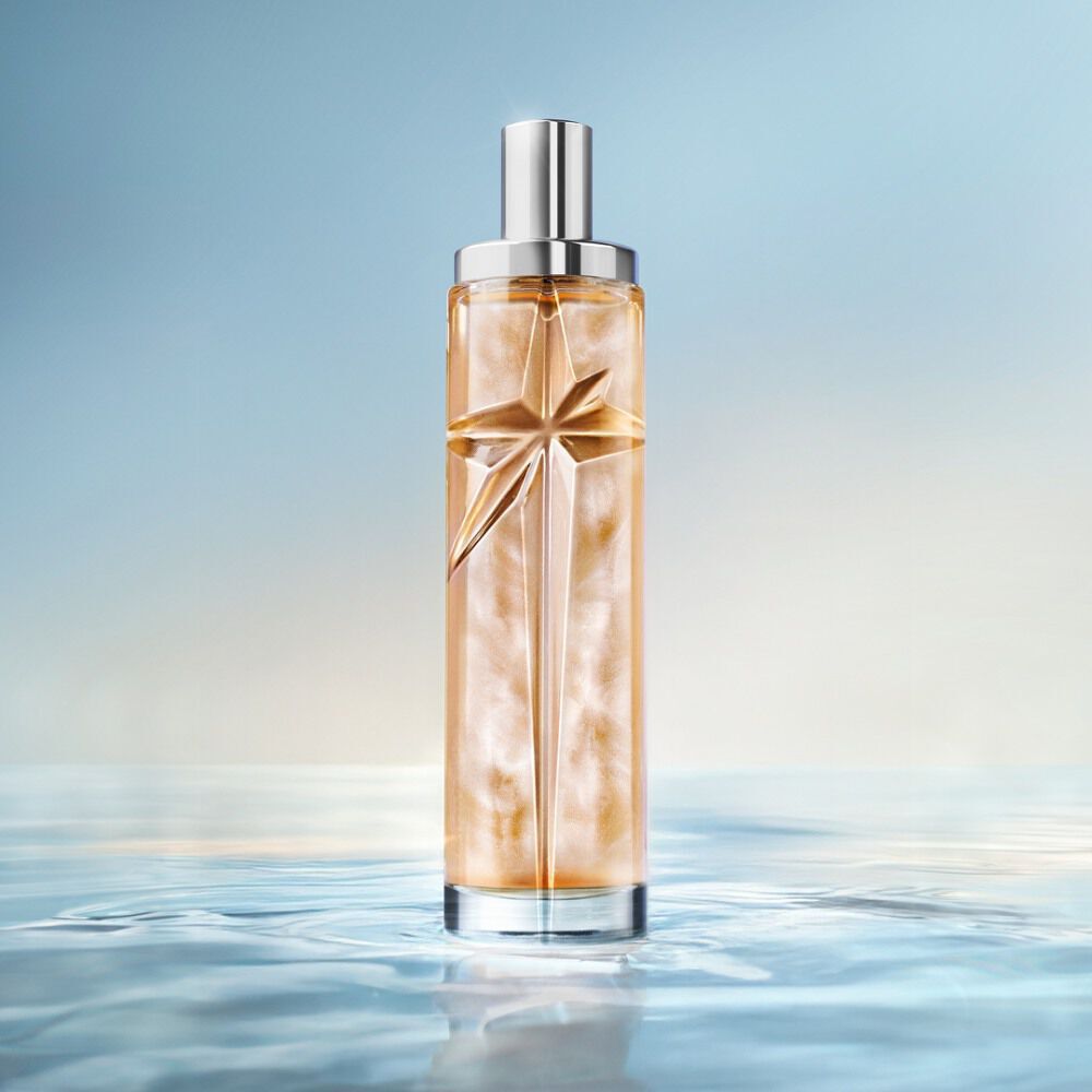 Elegant tall cylindrical perfume bottle with silver cap, filled with warm golden-amber juice, standing on a reflective surface mimicking calm water against a soft light blue background.