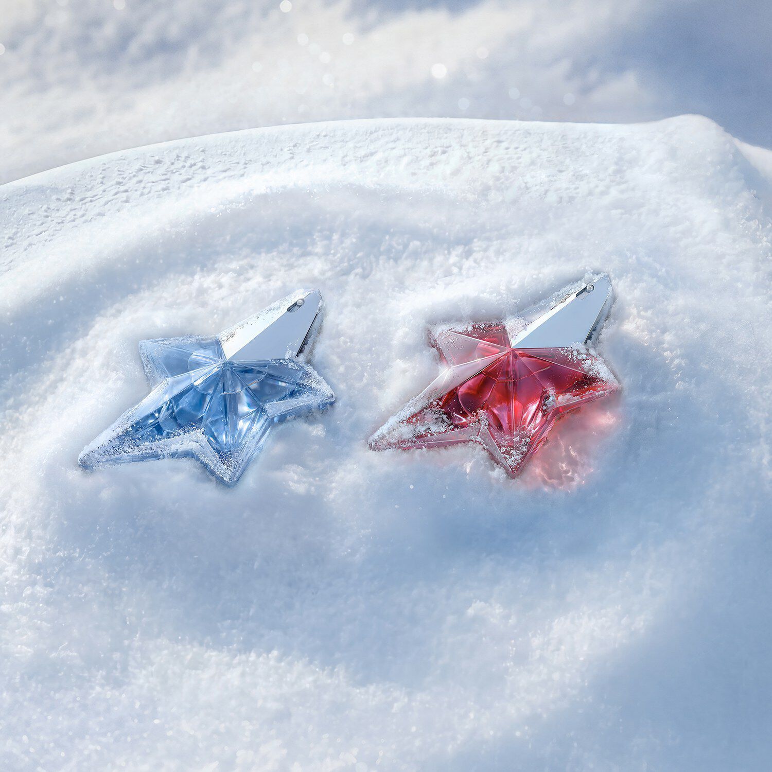 Two star-shaped Mugler perfume bottles—one pastel blue (Angel eau de parfum) and one bright pink (Angel Nova) —resting together in a soft drift of white snow.