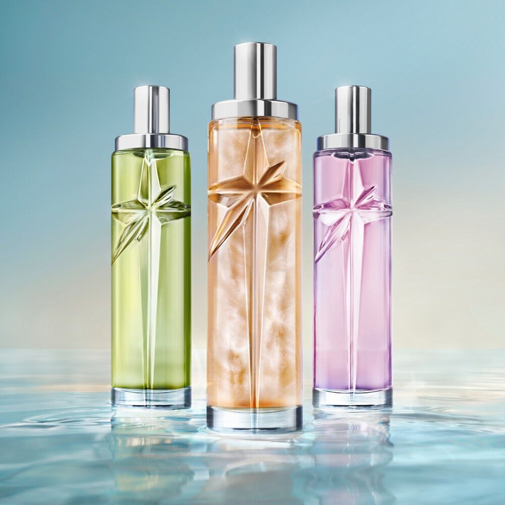The new Starlicious collection: three tall cylindrical perfume bottles with silver caps, in light green, golden-amber, and lavender-purple, standing side-by-side on a reflective water-like surface against a soft blue gradient.