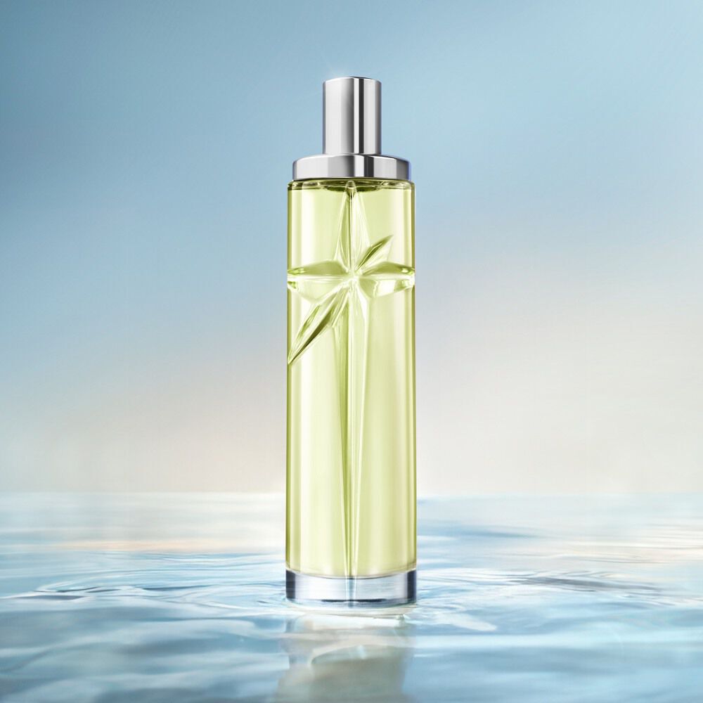 An elegant, tall, cylindrical perfume bottle with a silver cap stands vertically against a soft, light blue background. The bottle is filled with a pale green juice, and it sits on a reflective surface that mimics the ripples of clear, calm water, with the horizon line softly blurred in the background.