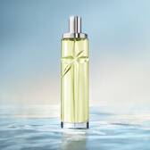 An elegant, tall, cylindrical perfume bottle with a silver cap stands vertically against a soft, light blue background. The bottle is filled with a pale green juice, and it sits on a reflective surface that mimics the ripples of clear, calm water, with the horizon line softly blurred in the background.