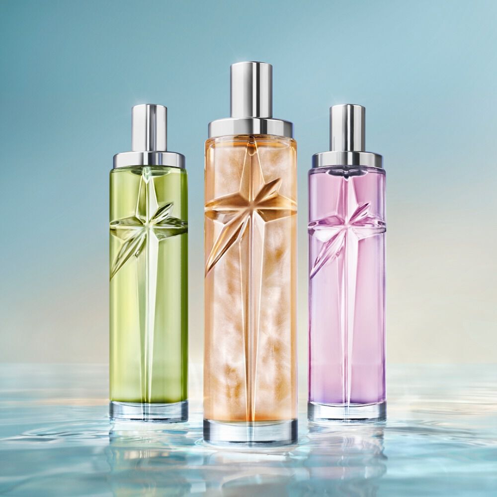 The new Starlicious collection: three tall cylindrical perfume bottles with silver caps, in light green, golden-amber, and lavender-purple, standing side-by-side on a reflective water-like surface against a soft blue gradient.