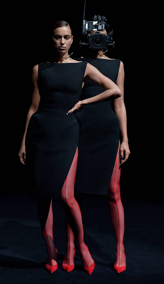 A model wearing a sleek black dress with high slits and red stockings stands confidently. The model's back is facing the camera, and a vintage camera is prominently placed where her head would be, creating a striking visual effect.