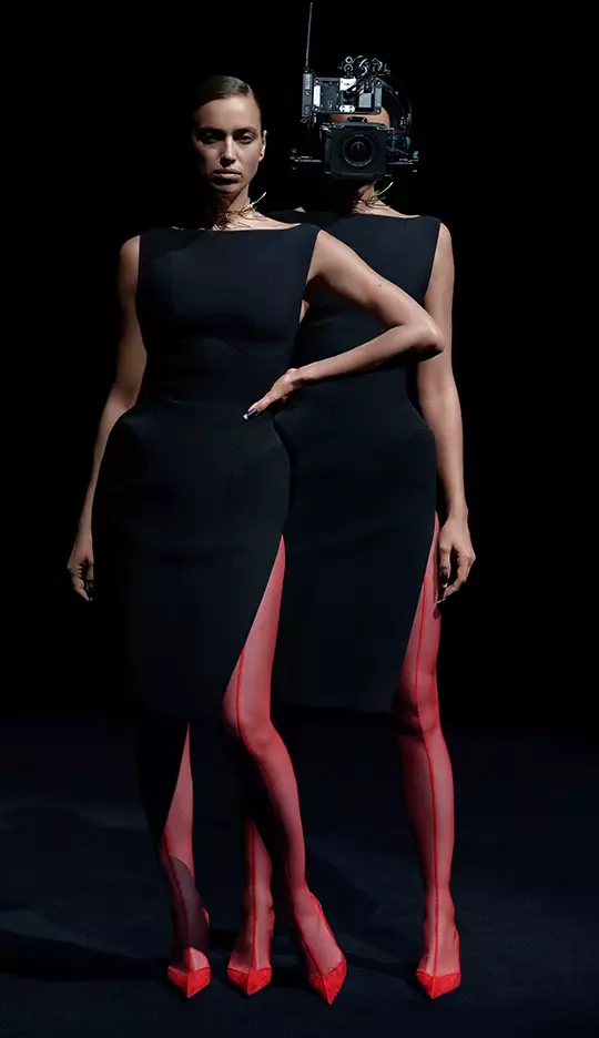 A model wearing a sleek black dress with high slits and red stockings stands confidently. The model's back is facing the camera, and a vintage camera is prominently placed where her head would be, creating a striking visual effect.
