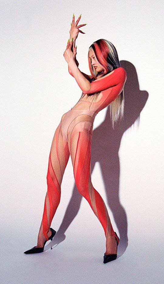 A figure poses dynamically in a striking body suit featuring a blend of nude and red colors, with bold striped patterns. The model has long, multi-toned hair and is wearing high-heeled shoes, casting an interesting shadow against a plain backdrop.
