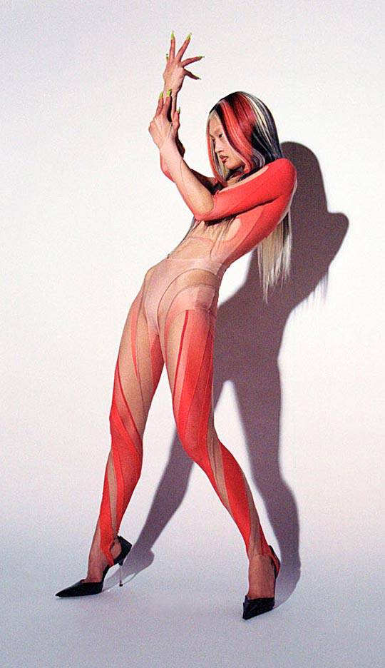 A figure poses dynamically in a striking body suit featuring a blend of nude and red colors, with bold striped patterns. The model has long, multi-toned hair and is wearing high-heeled shoes, casting an interesting shadow against a plain backdrop.