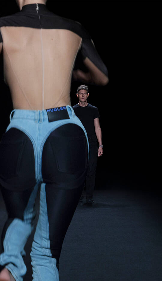 A model walks down a runway wearing a unique outfit featuring exaggerated denim and black fabric elements, while a man in a black shirt looks on from the distance. The scene is set against a dark background.