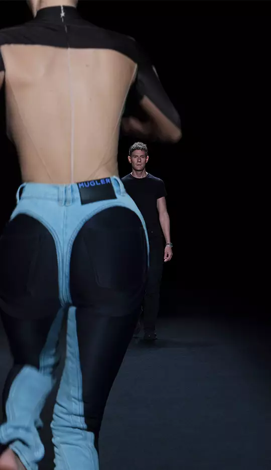A model walks down a runway wearing a unique outfit featuring exaggerated denim and black fabric elements, while a man in a black shirt looks on from the distance. The scene is set against a dark background.