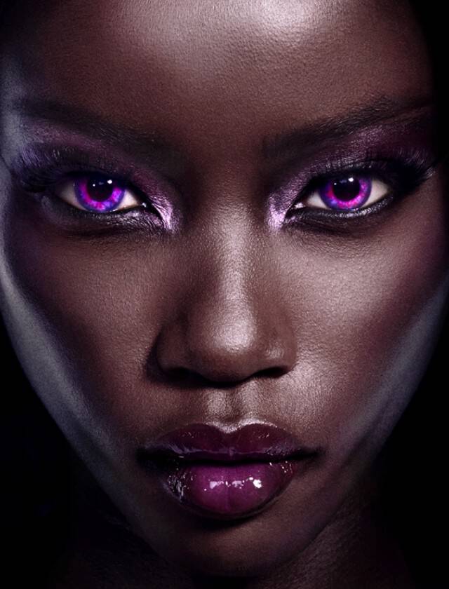A close-up of a model's face with striking purple eye makeup and glowing purple eyes against a dark background. The skin has a smooth, dark tone, adding to the dramatic effect.