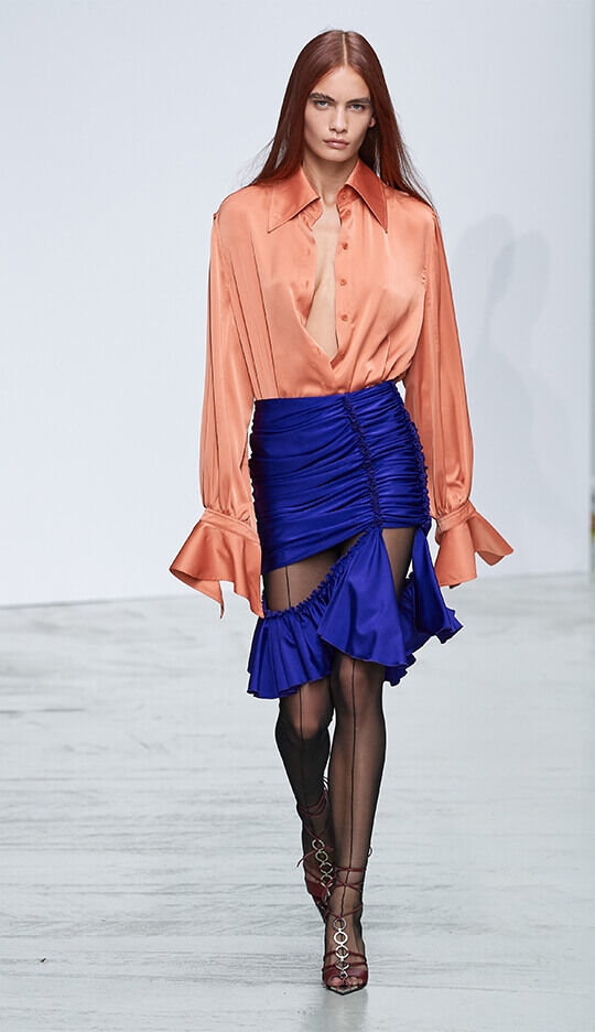 A model walks down the runway wearing an oversized orange silk shirt and a vibrant blue ruched mini skirt, paired with black sheer tights and strappy red heels. The background is a minimalist white display.
