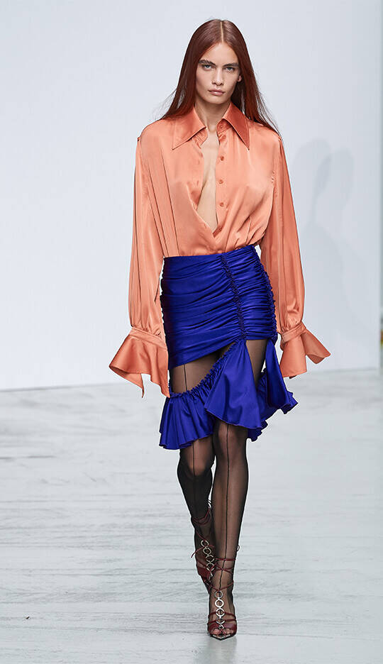 A model walks down the runway wearing an oversized orange silk shirt and a vibrant blue ruched mini skirt, paired with black sheer tights and strappy red heels. The background is a minimalist white display.