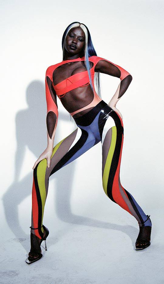 A model poses confidently in a vibrant, multi-colored outfit featuring bold cutouts, showcasing a combination of bright red, blue, yellow, and black against a minimal background.