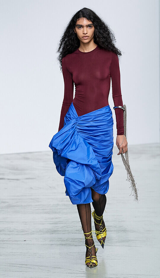 A model walks down the runway wearing a burgundy long-sleeve top and a voluminous blue skirt, paired with yellow strappy heels, showcasing a modern fashion design.
