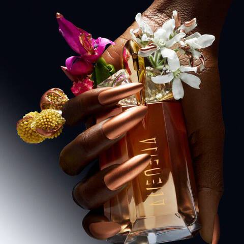 A hand adorned with vibrant rings holds a perfume bottle surrounded by colorful flowers against a gradient background.