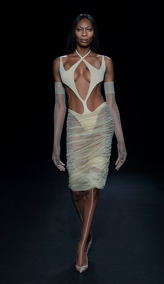 A model walks down the runway wearing a form-fitting, sheer dress with cut-out details, paired with matching gloves and high heels, against a dark background.