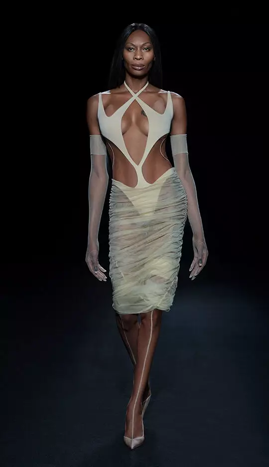 A model walks down the runway wearing a form-fitting, sheer dress with cut-out details, paired with matching gloves and high heels, against a dark background.