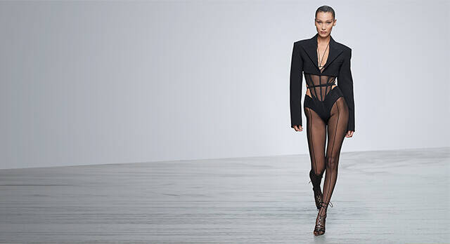 A model walks down a runway, showcasing a stylish black outfit with sheer panels and a structured blazer. The minimalist background emphasizes the fashion design.