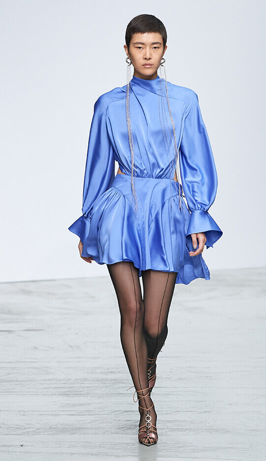 A model walks down a runway wearing a sleek, blue satin dress with a high neckline and dramatic sleeves, paired with sheer black tights and strappy heels. The background is minimalist and light.