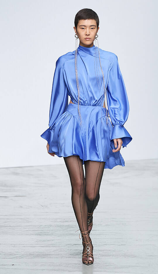 A model walks down a runway wearing a sleek, blue satin dress with a high neckline and dramatic sleeves, paired with sheer black tights and strappy heels. The background is minimalist and light.
