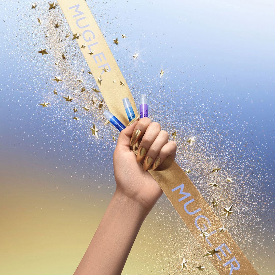 A hand with gold chrome nails grips a gold Mugler ribbon while holding several small fragrance sample vials; glitter and gold star confetti stream across a blue‑to‑gold gradient background.