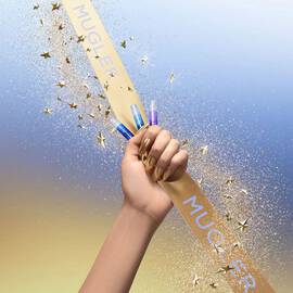 A hand with gold chrome nails grips a gold Mugler ribbon while holding several small fragrance sample vials; glitter and gold star confetti stream across a blue‑to‑gold gradient background.