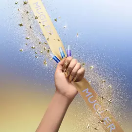 A hand with gold chrome nails grips a gold Mugler ribbon while holding several small fragrance sample vials; glitter and gold star confetti stream across a blue‑to‑gold gradient background.