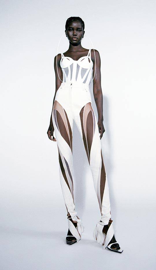 A model stands against a plain background, wearing a stylish, form-fitting white outfit with cut-out details and brown accents, paired with high-heeled shoes.