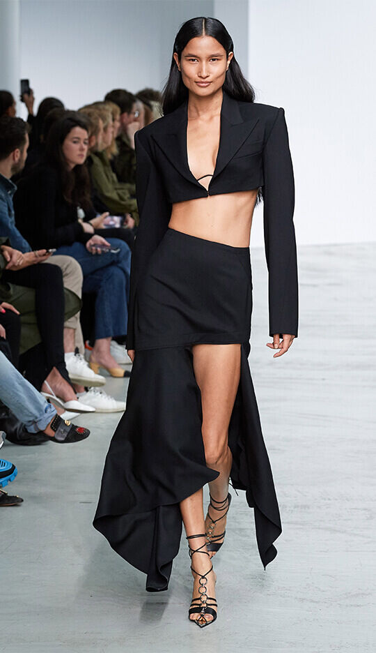 A model walks down a runway wearing a black cropped blazer and a high-slit skirt, showcasing a modern and bold fashion look. The audience in the background watches attentively.