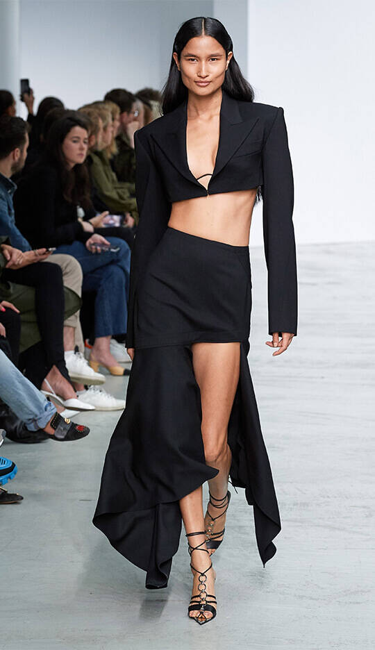 A model walks down a runway wearing a black cropped blazer and a high-slit skirt, showcasing a modern and bold fashion look. The audience in the background watches attentively.