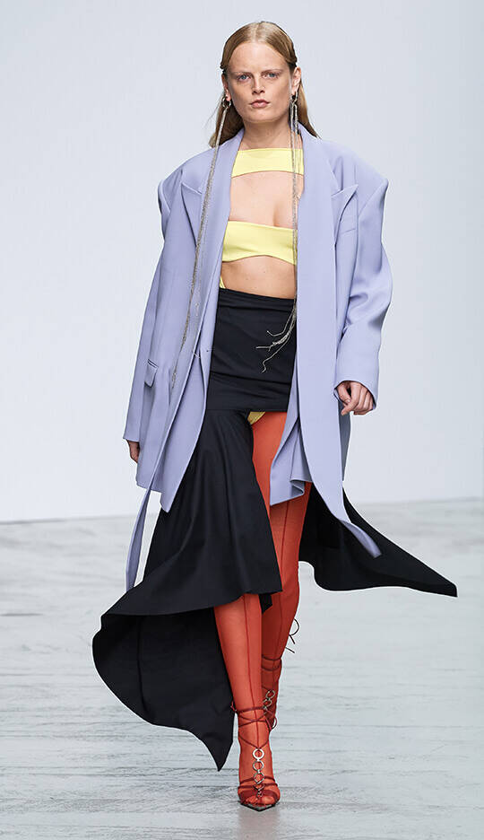A model walks down a runway wearing a layered outfit featuring a lavender oversized blazer, a yellow crop top, a black asymmetrical skirt, and orange tights, complemented by strappy red heels.