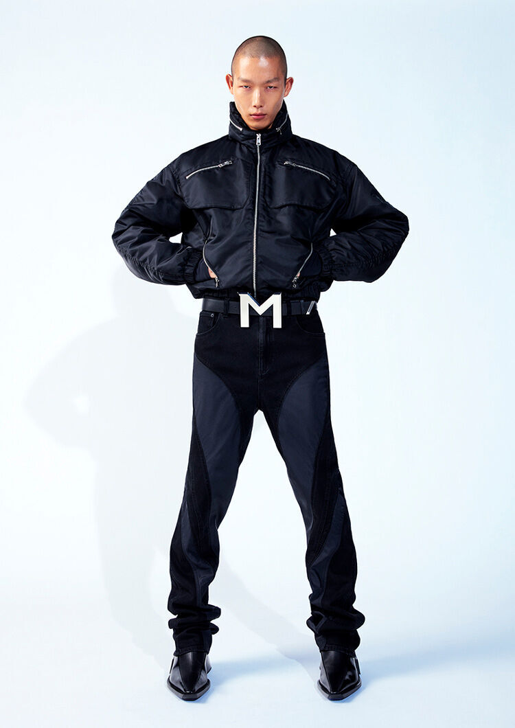 A model stands confidently, wearing a black zip-up jacket and black jeans with a noticeable belt featuring a large "M" buckle. The background is a soft, light blue, emphasizing the outfit's bold design.