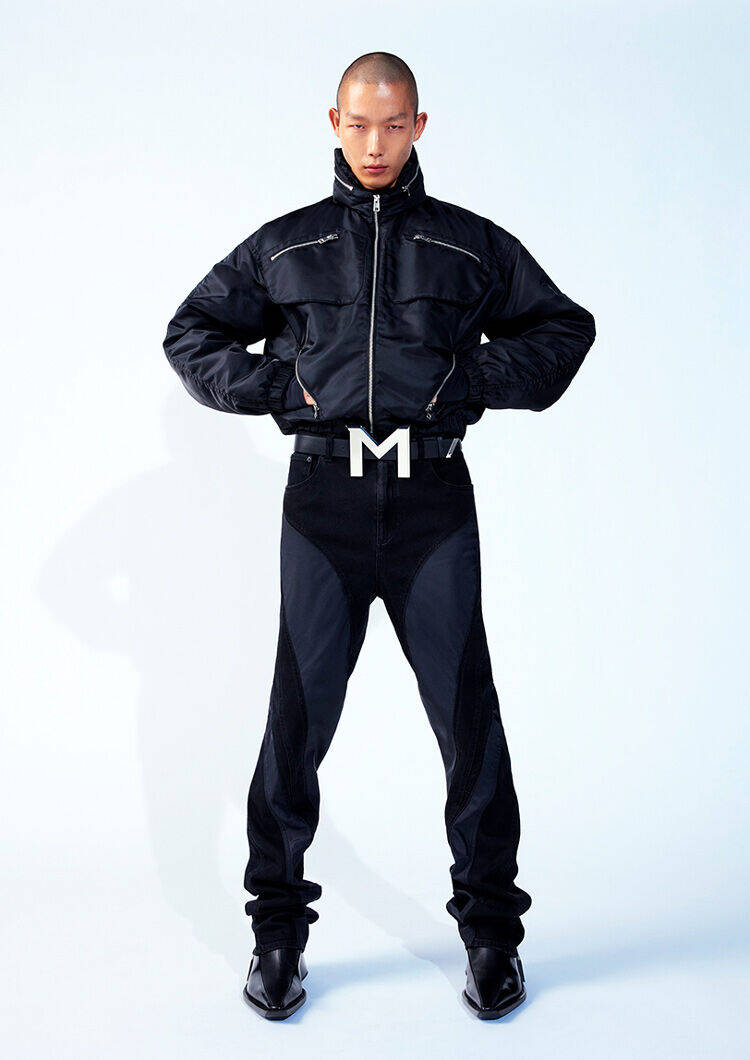 A model stands confidently, wearing a black zip-up jacket and black jeans with a noticeable belt featuring a large "M" buckle. The background is a soft, light blue, emphasizing the outfit's bold design.