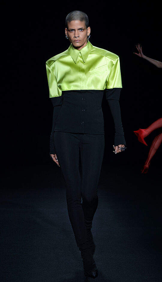 A model walks down the runway in a striking outfit featuring a bright green, oversized satin shirt with a fitted black base layer and sleek black pants, showcasing a modern and bold fashion statement.