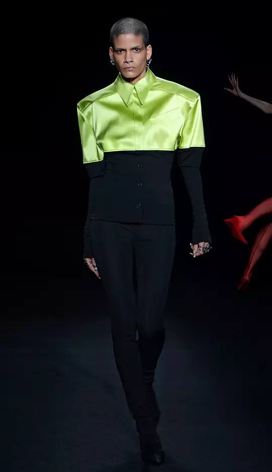 A model walks down the runway in a striking outfit featuring a bright green, oversized satin shirt with a fitted black base layer and sleek black pants, showcasing a modern and bold fashion statement.