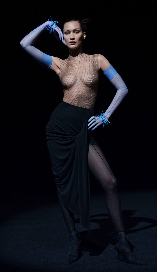 A model poses dramatically in a sheer, embellished bodysuit and a form-fitting black skirt, with striking blue gloves and a confident expression. The background is dark, emphasizing her silhouette and the outfit's details.