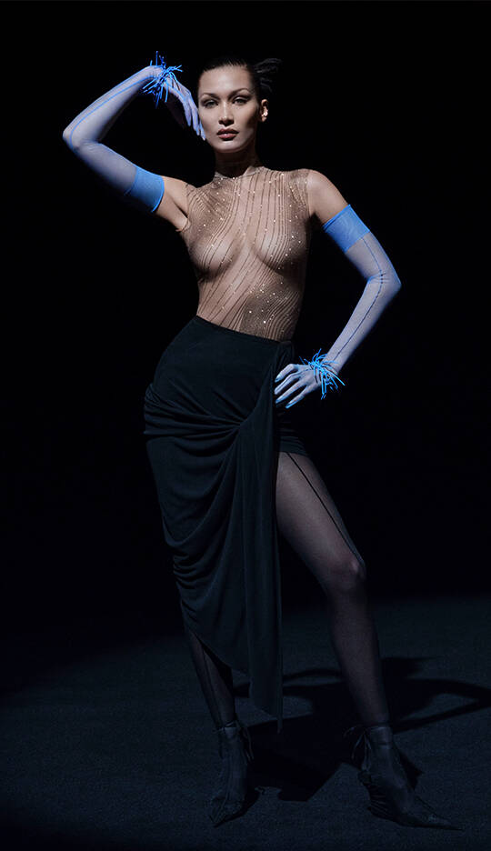 A model poses dramatically in a sheer, embellished bodysuit and a form-fitting black skirt, with striking blue gloves and a confident expression. The background is dark, emphasizing her silhouette and the outfit's details.