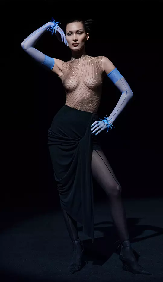 A model poses dramatically in a sheer, embellished bodysuit and a form-fitting black skirt, with striking blue gloves and a confident expression. The background is dark, emphasizing her silhouette and the outfit's details.