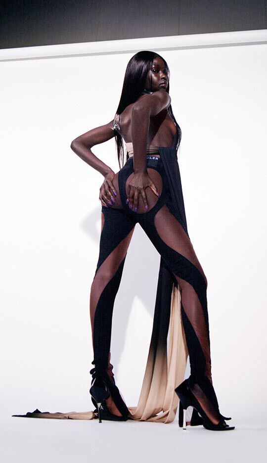 A model poses dramatically in a stylish black outfit with cutouts, showcasing bold fashion against a minimalist backdrop. The ensemble features sheer fabric and high heels, highlighting a striking silhouette.