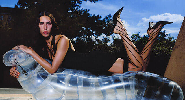A model in a black dress lounges on a futuristic, transparent couch, wearing high-heeled shoes. The background features a bright blue sky with clouds and greenery.