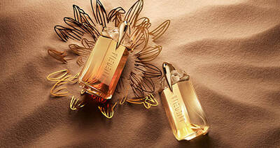 Two elegant perfume bottles are positioned atop a textured sand surface, surrounded by delicate golden floral outlines. The bottles feature a sleek, modern design with varying shades of amber and gold.