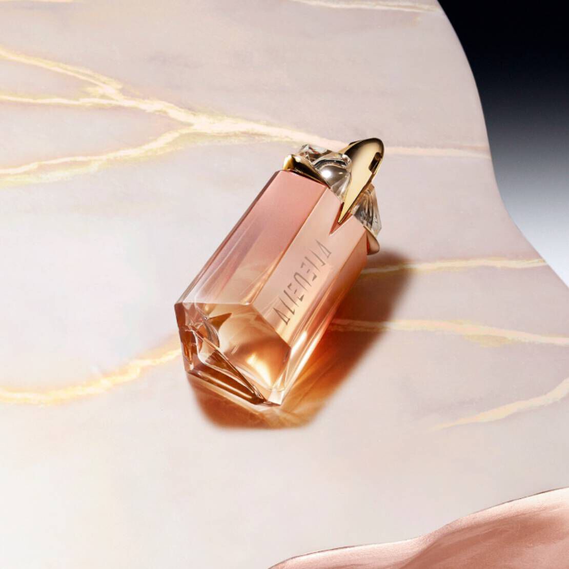 A stylish glass perfume bottle with a rose-gold hue sits elegantly on a smooth, light-colored surface, featuring unique geometric design and a decorative cap.