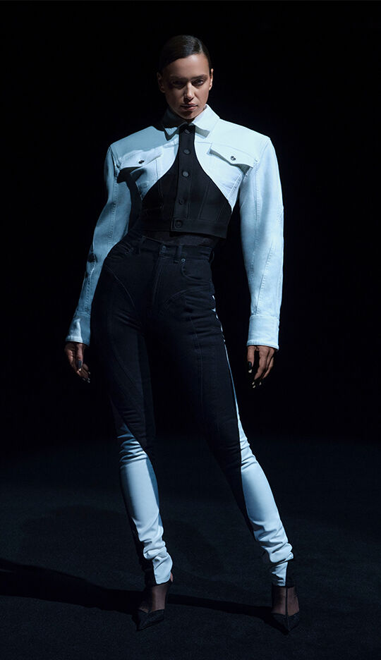 A model poses in a dramatic fashion outfit featuring a two-tone jacket and fitted pants, set against a dark background.