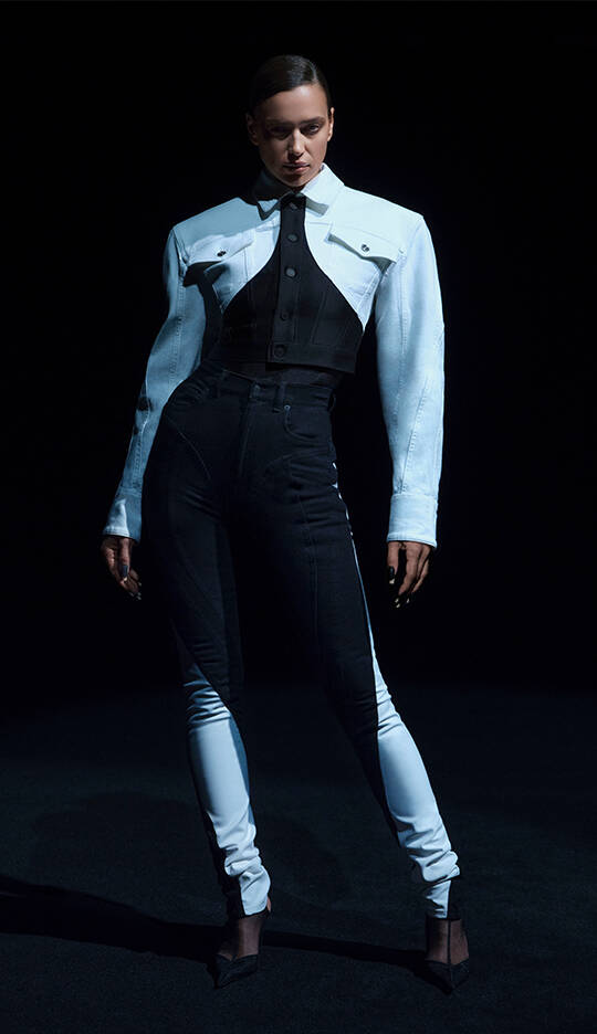 A model poses in a dramatic fashion outfit featuring a two-tone jacket and fitted pants, set against a dark background.