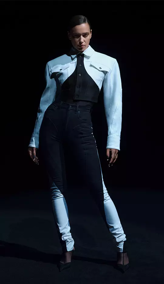 A model poses in a dramatic fashion outfit featuring a two-tone jacket and fitted pants, set against a dark background.