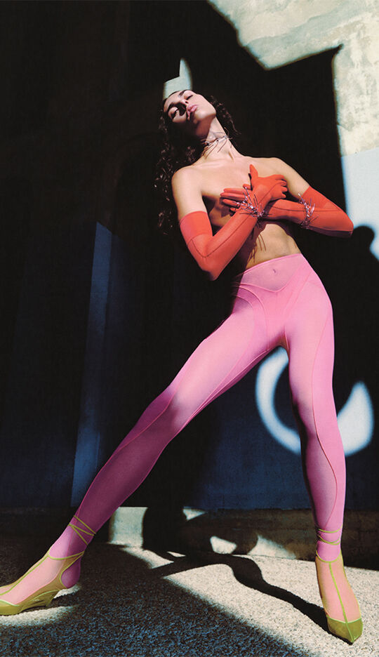 A model poses dramatically in a vibrant outfit featuring a crop top and gradient legging that transition from pink to purple. She wears long orange gloves and striking yellow strappy heels, with a bold shadow cast against a textured wall behind her.