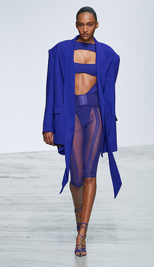 A model walks down the runway wearing a striking blue ensemble that includes a cropped top, high-waisted sheer pants, and an oversized blazer. The outfit features bold lines and a modern silhouette, complemented by stylish sandals.