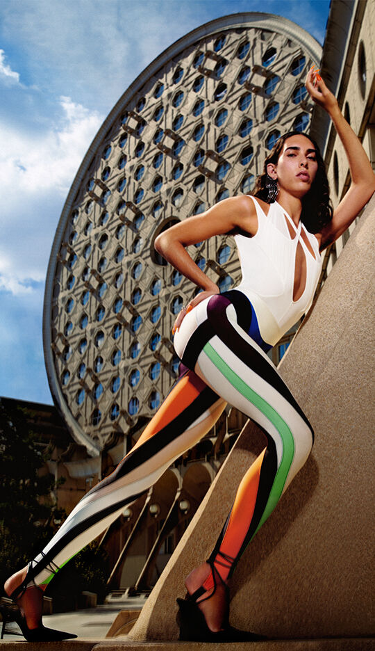 A model poses dramatically against a modern architectural backdrop, wearing colorful, patterned leggings and a stylish white top. The scene features a vibrant blue sky and geometric shapes in the architecture.