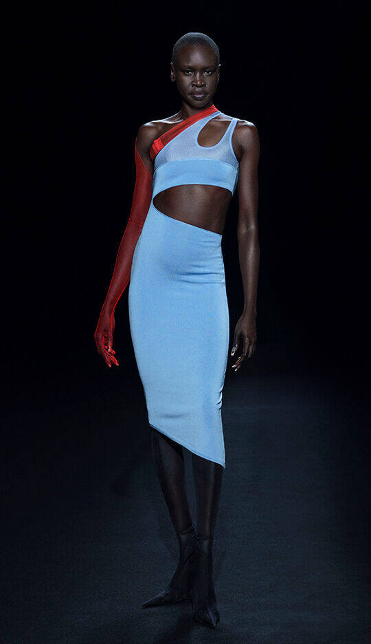 A model walks the runway in a striking blue form-fitting dress with a unique cut, paired with a long red glove on one arm, showcasing a bold and modern fashion design against a dark background.