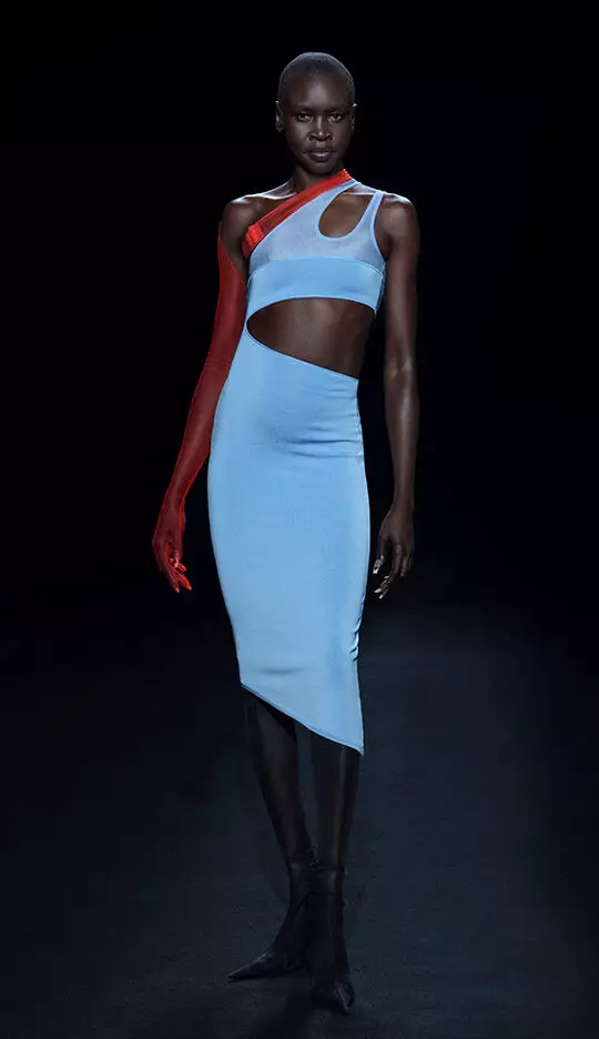 A model walks the runway in a striking blue form-fitting dress with a unique cut, paired with a long red glove on one arm, showcasing a bold and modern fashion design against a dark background.