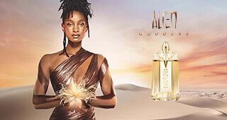 A woman in a shimmering bronze outfit stands confidently in a desert landscape during sunset, holding a feather-like accent. Beside her is a bottle of the "Alien Goddess" perfume.